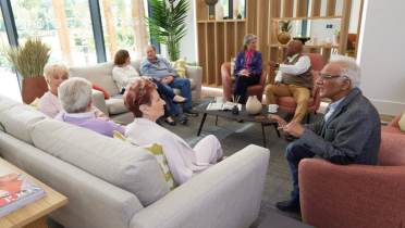 A group of elderly people sit and chat in a modern, well-lit lounge area with sofas, armchairs, and a coffee table.