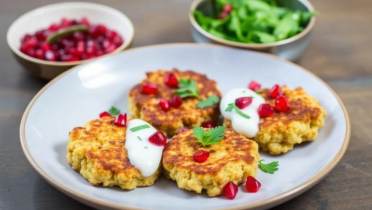 Three golden fritters topped with white sauce, pomegranate seeds, and parsley are served on a plate, with bowls of salad and pomegranate seeds in the background.