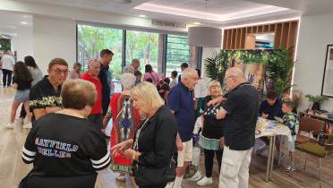 A group of people, mostly seniors, gather and socialize indoors. Some are seated at a table, while others stand and converse in a bright, modern room.
