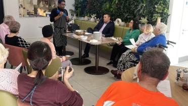 A chef gives a cooking demonstration to a seated group of adults in a modern room with a green plant wall.